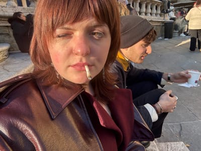 The image is a close-up, selfie-style photograph capturing a young woman and individuals in the background within Piazza Grande, Arezzo, Italy. The primary subject, a woman with reddish-brown hair and bangs, wears a dark reddish-brown leather-look jacket. A hand-rolled cigarette, consistent in appearance with cannabis, is held between her lips with a visible ember; her left eye is closed or squinting. She has a nose ring on her left nostril.
Seated on stone steps to her right and slightly behind is a young man with dark hair and a beard, wearing a dark knit beanie and a dark jacket over a yellow shirt. He holds a white disposable cup in his left hand and a smartphone in his right, looking away from the camera. A silver watch and bracelet adorn his left wrist. Discarded cigarette butts lie on the steps near him.
In the left background, another woman in a dark, fur-hooded winter coat is visible, seated. Additional indistinct individuals are present in the far background on the stone steps and pavement, some emitting visible smoke. Architectural elements include a stone balustrade with small columns and a black trash receptacle. The scene is outdoors, illuminated by strong, warm sunlight, casting visible shadows.