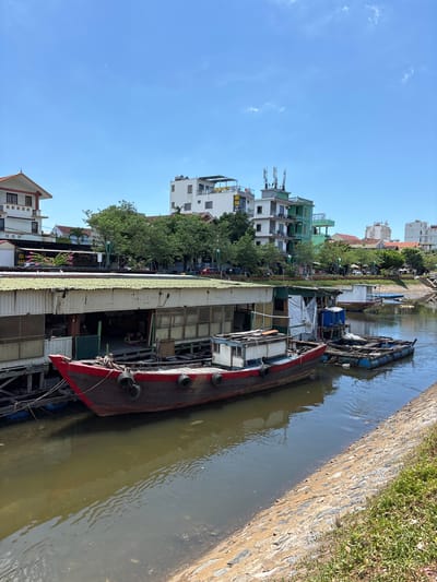 This is a vertically oriented, eye-level, medium shot of a canal in Đồng Hới Ward, Vietnam, on a clear, sunny day. The canal runs horizontally across the frame from right to left. Along the left bank of the canal are several buildings, including a long, single-story structure with a green corrugated metal roof and large openings. In front of this structure, a dark red and brown wooden boat with black fenders is moored. Further to the right, a smaller boat with blue barrels is tied to the larger one. The right bank of the canal is lined with a stone embankment covered in green grass. In the background, various multi-story buildings and trees are visible, with a clear blue sky overhead. No people are visible in the image.