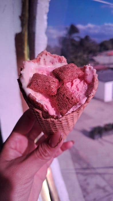 A close-up captures a hand holding an ice cream cone. The cone is filled with pink ice cream and topped with small, square, dark brown cereal pieces. The waffle cone has a textured, crisscross pattern. The background is blurry but appears to include both indoor and outdoor elements with blue sky. The overall lighting gives the scene a slightly pink hue.