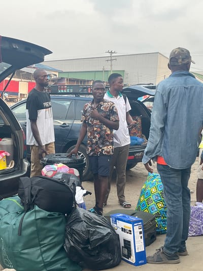 Four men load luggage into the trunks of two dark cars. The luggage includes backpacks, duffel bags, and a spare tire. One man wears a denim jacket and camouflage hat. A man in a white shirt stands next to a man wearing a patterned shirt and blue shorts. The fourth man wears a white t-shirt with black sleeves.
