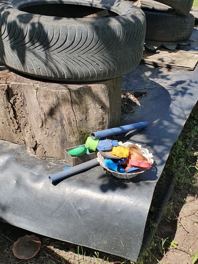 In Novoivanivka, Ukraine, a close-up, eye-level outdoor shot shows a collection of repurposed tires and gardening tools on a black plastic tarp. The foreground features a light-colored, lacy basket filled with small, colorful plastic scoops and molds in shades of blue, yellow, and red. Beside the basket, two dark blue plastic pipes lie horizontally, one in front of the other. To the left of the pipes, a small, rectangular metal container holds a bright green plastic scoop and a smaller blue scoop. A large, weathered wooden stump serves as a base for a portion of a black tire, which is partially obscured by shadows cast by unseen foliage. The tire has a distinct tread pattern. In the background, another tire rests on wooden planks. The ground surrounding the tarp is dry earth with sparse green vegetation and some scattered debris, including a dark, mushroom-like object near the bottom left corner of the frame. The scene appears to be a DIY garden or play area, possibly in a rural or suburban setting.