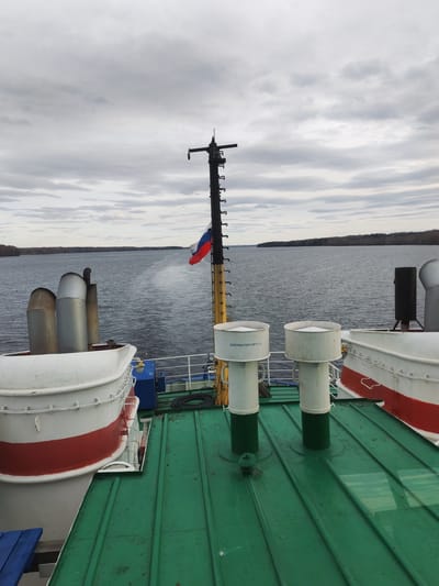 The image depicts a view from the deck of a vessel on a body of water. In the background, land is visible, potentially indicating the shoreline. The deck is green and features various shipboard elements, including white lifeboats with red striping, cylindrical vents, and a mast with the Russian flag. The sky is overcast with grey clouds. The location is in the city of Yakunina Gora, Russia.