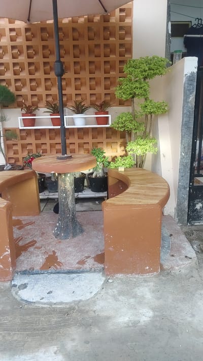 A curved bench and round table made of concrete and wood are situated outdoors in a yard, likely in Setiamulya, Indonesia. A partial umbrella shades the scene. The area is decorated with potted plants and a patterned wall. The ground is made of concrete with some water stains.