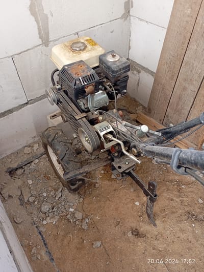 The image shows a single-wheel, engine-powered tiller or cultivator. The tiller is black with a yellow fuel tank and has a ribbed belt drive connected to the engine. It is parked on a dirt and gravel surface, with some scattered debris. Behind the tiller is a wall constructed of cinder blocks, with some unfinished concrete work. To the right of the tiller are several wooden planks leaning against the wall. The scene appears to be an outdoor or semi-outdoor storage area, possibly a shed or an uncompleted structure. The lighting suggests it is daytime. A timestamp on the image reads "20.06.2026 17:52", indicating the date and time the photo was taken. The location is specified as Урыцкі сельскі Савет, Belarus. There are no people visible in the image.