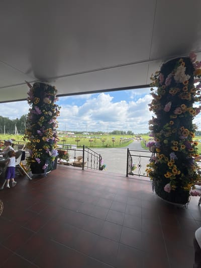 The image depicts an outdoor seating area with a tiled floor, likely at a restaurant or cafe, in Akhlebaikha (Kovriginskii S/S), Russia. Two large, cylindrical floral arrangements, adorned with sunflowers and other artificial flowers, stand prominently near the entrance to an outdoor pathway. In the foreground, a person wearing a white cap and a child in purple shorts and a white top are seated at a table. The pathway leads to a green field with scattered trees and a few parked cars visible in the distance under a partly cloudy sky. A black metal railing borders the steps to the pathway.