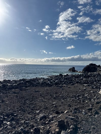 The image displays a sunlit volcanic coastline in Santiago del Teide, Spain. The foreground is dominated by a dark beach comprised of numerous irregular black and dark grey volcanic rocks, pebbles, and coarse sand. The mid-ground features a calm, deep blue ocean, reflecting intense sunlight on its left side. Several rugged, dark volcanic rock formations emerge from the water and line the right side of the shoreline. The upper portion of the scene reveals a bright blue sky with scattered white cumulus clouds, more concentrated along the distant horizon. A prominent sun flare originates from the upper left quadrant of the image. No individuals, structures, or artificial objects are present.