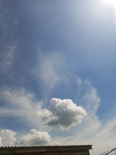 A bright, sunny day is depicted in Zavyalovo, Russia, with a clear blue sky dominating the frame. Wispy, white cirrus clouds are scattered across the upper half of the image, with a larger, more voluminous cumulus cloud situated in the lower center. The sun is visible in the upper right corner, creating a bright glare. Along the bottom edge of the image, a corrugated roof of a structure, likely a shed or a low-rise building, is visible, made of what appears to be dark wood panels. There are no people or animals visible in the image. Electrical wires are present on the far right side of the image, running diagonally upwards.