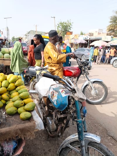 A bustling outdoor scene showcases a street in an urban African setting. Stacks of mangoes are displayed near a motorcycle, with a man in a golden outfit sitting on another nearby. A woman with dark hair stands close by, while others are gathered in the background. A street corner with shops and cars, including a taxi, completes the vivid picture of daily life in the town.