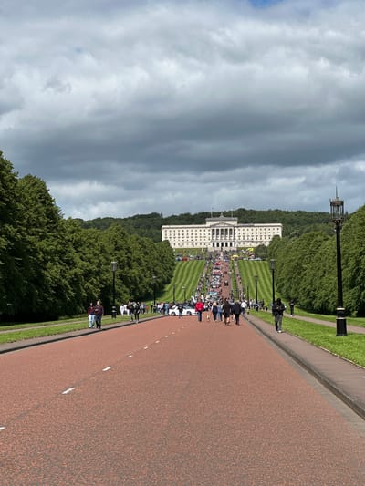The image shows a long, sloping pathway leading towards a large white building with classical architecture, identified as Stormont, the Parliament Buildings of Northern Ireland, located in Belfast, United Kingdom. The pathway is lined with mature trees on either side, and the grassy slope in front of the building is striped with mown grass. A crowd of people are walking up the pathway and gathered on the slope, with a number of cars parked on the grass. The sky is overcast with dramatic dark clouds. The foreground is dominated by the red-toned tarmac of the pathway with white dashed lines indicating lanes. Several street lamps are visible along the pathway. The scene suggests a public event or gathering at the Parliament Buildings.