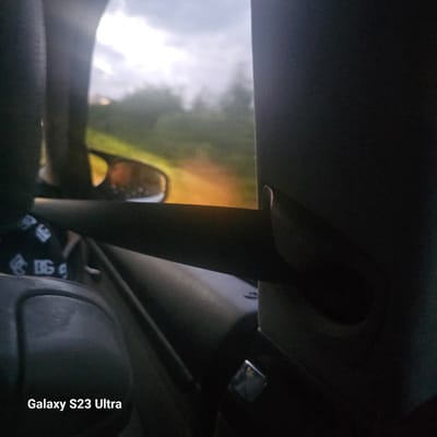 The image shows a view from inside a vehicle, looking out the window. The exterior scene is blurred, with green foliage and a dirt road visible. A side-view mirror reflects an orange-red object. The interior of the car is in focus in the foreground, with a dark seat and a black strap with white logos on it. The bottom left corner of the image displays the text "Galaxy S23 Ultra". The image does not provide any information about the city of Manastir, Bulgaria.