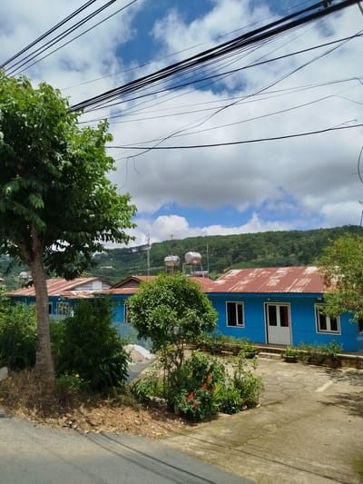 In Xuân Sơn, Vietnam, a bright blue building with a weathered orange metal roof is visible under a cloudy sky. Several solar water heaters are mounted on the roof. The building is surrounded by lush green vegetation, including a large tree on the left side of the frame. The foreground shows a paved area and a road, suggesting a residential or commercial property. In the background, rolling hills covered in dense green trees rise up, with some structures and a communication tower visible on the slopes. Power lines crisscross the sky above the scene.
