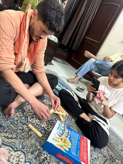 The image shows three individuals sitting on a rug, possibly in a home in Zirakpur, India. A man in an orange scarf is building a structure with wooden planks, the "brain builder" wooden building planks, in front of him. A girl is looking at a cell phone. An additional person is partially visible, sitting behind the girl. The rug is patterned.