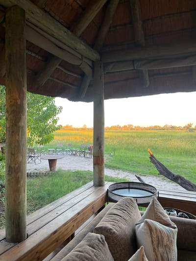 The image depicts a scene from a lodge or similar structure in Khwai, Botswana. It is viewed from an elevated, partially enclosed wooden deck with a thatched roof supported by wooden beams. Outside, a grassy plain stretches to the horizon under a yellow-tinged sky, possibly during sunrise or sunset. Seating, including a circular table and chairs, are visible in the distance. A tree is present in the foreground, partially obstructing the view. The deck includes a cushioned sofa with pillows, a round metal table, and wooden planks forming the floor and railing.
