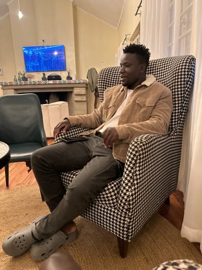 An indoor scene features an adult male seated in a high-backed armchair upholstered in a black and white houndstooth pattern. The man, with dark skin and short textured hair, wears a light brown corduroy jacket with a fleece collar over a light-colored ribbed shirt, dark gray cargo-style pants, and light gray Crocs-style slip-on shoes. He is positioned with his torso angled slightly to the right, his left arm resting on his thigh where a dark object, possibly a smartphone or remote, is visible. His legs are crossed at the ankles.
To the left of the man, a dark green upholstered chair and a portion of a round, dark-surfaced table are visible. In the background, a stone fireplace structure with a mantelpiece spans the wall. A flat-screen television is mounted above the mantelpiece, displaying a scene with multiple individuals, possibly a performance, under blue lighting. The mantelpiece itself holds several decorative items including a JBL portable speaker, various vases and bottles, and two dark figurines. A light-colored, possibly off-white, upholstered stool is partially visible to the left of the fireplace opening.
White sheer curtains cover a window on the right side of the frame. The floor beneath the armchair is covered by a woven natural fiber rug. The walls are light-colored, and a single bare bulb light fixture hangs from the ceiling in the upper left. The specific location within Olkaria ward, Kenya, is not discernible from the visual evidence provided.