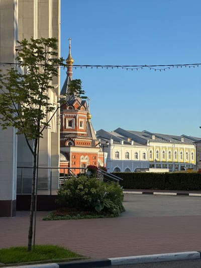 The image shows a view of a street in Yazvitsevo, Russia, featuring a bright blue sky and sunlight illuminating various buildings. In the foreground, a modern building's light-colored facade is visible on the left, with a young tree and green grass growing from a square patch of ground. A metal railing separates this area from a paved walkway and a landscaped planter box filled with lush green bushes and red flowers.
Dominating the mid-ground is a vibrant red brick church with intricate white detailing, a dark grey roof, and a golden dome topped with a cross. To the right of the church, a row of long, light yellow and cream-colored buildings with arched windows and grey roofs stretches into the distance. A dark green hedge runs along the front of these buildings. A string of decorative lights is suspended across the sky. The ground is paved with reddish-brown bricks. No people are visible in the scene.