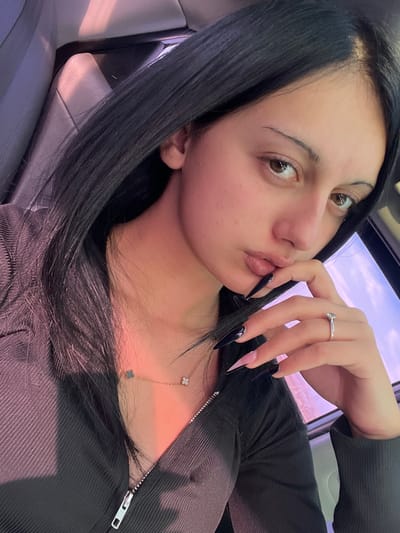 A female individual is depicted in a close-up shot, seated within the interior of a vehicle. Her dark, straight hair is visible, falling over her shoulders. She wears a dark-colored, possibly ribbed, long-sleeved top with a partially unzipped front. A gold-colored necklace with two clover-shaped charms (one dark, one light) is visible around her neck. Her right hand is raised, with fingers positioned near her mouth, displaying long, pointed artificial nails predominantly dark in color with a pink gradient near the base of some nails. A silver-colored ring with a single clear stone is worn on the ring finger of her right hand. Her lips are slightly pursed, and her eyes are directed towards the camera. The background shows a light-colored car seat with a headrest, and through a window, a light-colored sky with a faint pink hue. The specific location within the city of Surami, Georgia, cannot be determined from the visual information provided, as the scene is entirely confined to the vehicle's interior.