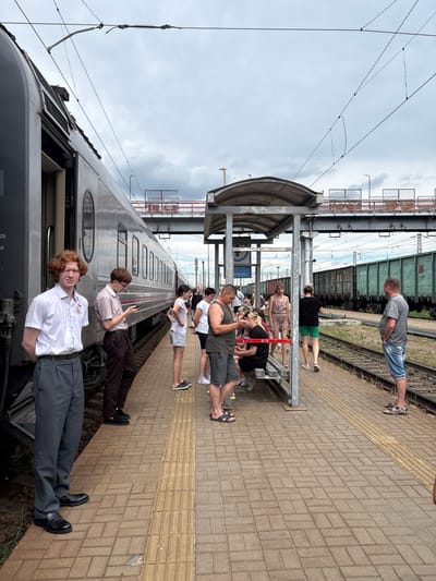 The image depicts a train station platform in Povorino, Russia, under an overcast sky. A grey passenger train is parked on the left, with its doors open, and several individuals are standing on the platform. To the right of the train, a covered waiting area with benches is visible, occupied by a few people. Beyond the platform, railway tracks lead to a line of green freight cars. The platform is paved with brickwork, and a tactile warning strip runs along the edge. Overhead, complex electrical wiring for the train is suspended. The overall atmosphere suggests a paused moment at a transportation hub.