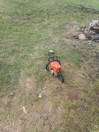 A gas-powered chainsaw, predominantly orange and black, lies on a patch of dry, uneven grass. A yellow and black measuring tape is attached to the top of the chainsaw's guide bar. To the right of the chainsaw, a pile of discarded building materials, including bricks, wood planks, and metal rebar, is visible, along with some scattered branches and stones. The background consists of more dry grass and a sparsely vegetated area. The scene appears to be in an outdoor, possibly rural, location in Удзелаўскі сельскі Савет, Belarus, suggesting a work or storage area.