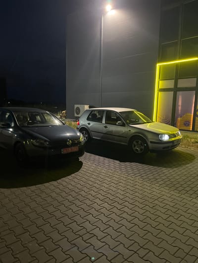 The image shows a dark, exterior scene at night, likely in Strumivka, Ukraine, with two Volkswagen Golf cars parked on a paved surface. A bright floodlight illuminates the area from the top left. The building behind the cars has a dark, corrugated metal facade and features a large glass window or door with a vibrant yellow neon light outline. To the right of the cars, an outdoor air conditioning unit is visible on the ground.
The first car, a dark colored Volkswagen Golf, is on the left. Its front license plate is visible and reads "03 B 7541". The headlights are off. Through the windshield, two figures can be seen seated in the front.
The second car, a silver Volkswagen Golf, is to the right of the first car. Its headlights are on, casting a bright beam. Its license plate is partially visible but not fully legible. The car is positioned at an angle, showcasing its side profile and front end.
The ground is paved with interlocking concrete blocks, arranged in a herringbone pattern. The overall lighting suggests it is nighttime, with artificial light sources dominating the scene. The sky is dark and appears clear.
