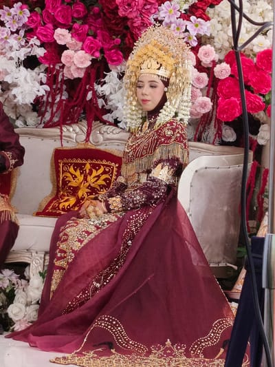 In Meuliek, Indonesia, a young woman is seated on a plush sofa, adorned in a traditional maroon wedding gown and an elaborate gold headdress. The backdrop is a vibrant floral arrangement of pink and white roses, accented with deep red tassels. The woman's attire features intricate gold embroidery and embellishments, with a matching maroon hijab and a flowing veil. A decorative maroon cushion with gold leaf patterns rests beside her. The setting appears to be a formal event, possibly a wedding ceremony or reception, given the celebratory attire and floral decorations.