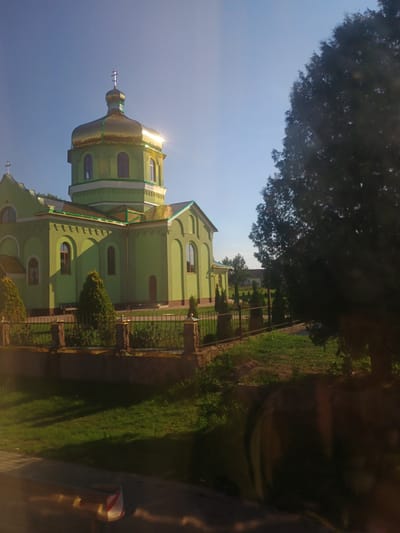 This media file depicts a sunlit scene featuring a prominent green church with a golden dome. The church is partially obscured by a large tree in the foreground, with its branches creating a dappled shadow on the building. The sky is a clear, bright blue, suggesting a sunny day. The church's architecture includes arched windows and decorative trim. A metal fence runs along the side of the church, separating it from a grassy area. The image appears to be taken outdoors, likely in a rural or semi-rural setting, given the presence of the tree and open sky. The orientation of the image is portrait, with the church occupying the majority of the frame. The sunlight glints off the golden dome, indicating it's likely daytime, possibly mid-morning or afternoon. No people are visible in the image, and no text is present. The overall impression is one of a peaceful, serene religious building on a pleasant day.