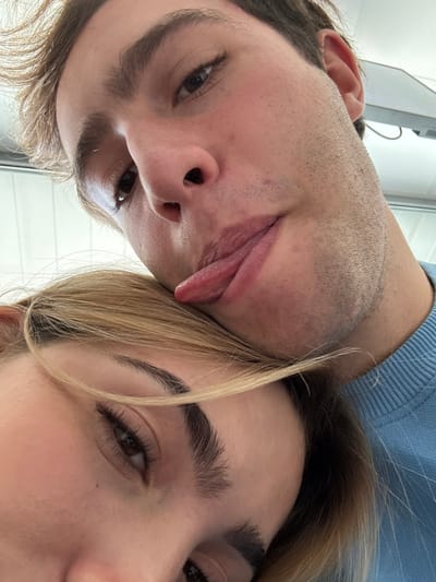 This close-up selfie depicts two individuals in Saint-Priest, France. The upper subject is a light-skinned male with short, light brown hair and visible stubble on his cheeks and chin. He wears a light blue crew-neck sweater and is seen looking towards the camera with his tongue extended from his mouth, resting his head on the lower subject's head. His right eye, brown in color, is visible. The lower subject is a light-skinned female with blonde hair that partially covers her face. Her right eye, brown with prominent eyelashes, and a well-defined eyebrow are visible. The background is uniformly light-colored and out of focus, suggesting an indoor setting, with a blurred rectangular object in the upper right corner.