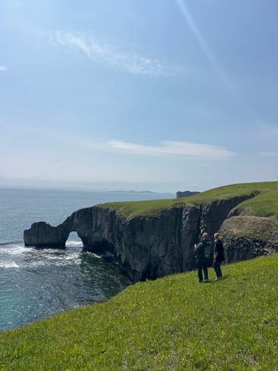 The image depicts a coastal scene with a rocky cliff face and grassy terrain at the top. Two people are standing on the grassy edge, seemingly observing the landscape. The cliff has a natural arch formation at its base, where the water from the ocean flows through. The sky is blue with some wispy clouds. The image does not depict the city of Slavyanka, Russia.