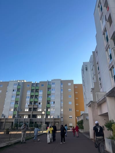 This image captures an urban residential scene in Noisy-le-Grand, France, on a bright, clear day. The primary subjects are several multi-story apartment buildings and a group of people outdoors.
On the left side of the frame, a tall, modern apartment building dominates, characterized by numerous windows and balconies, many of which feature distinctive green panels. In the background and to the right, other apartment blocks are visible, some with beige and orange facades, indicating a large residential complex. The sky is a vibrant, cloudless blue, and strong sunlight casts clear shadows on the buildings and ground, suggesting it is daytime.
Several individuals are present on a paved ground-level area in front of the buildings. In the foreground on the right, a person is seen from behind, wearing a black t-shirt with the text "VINI JR. 7" prominently displayed on the back, along with a white cap. Further down the path, a group of people, including some in red and pink attire, are gathered. Another individual in a light blue top is standing near a black and white soccer ball, suggesting a casual, recreational atmosphere. Other figures are scattered throughout the scene, either standing or walking along the path. Green foliage and bushes are visible near the bases of some buildings. The overall impression is a typical day in a residential neighborhood.