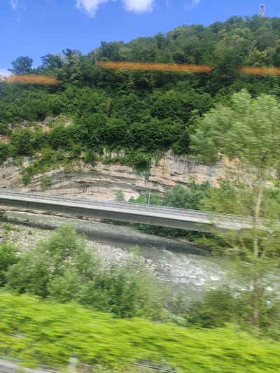 The image captures a scenic view of a river flowing through a valley, bordered by a steep, rocky cliff face covered in lush green foliage. A modern, multi-lane bridge spans across the river, its grey concrete structure and metal railings clearly visible. The surrounding environment is dominated by dense trees and vegetation, with a bright blue sky with scattered clouds above. In the distance, atop a hill, a radio tower is visible. The perspective suggests the photograph was taken from a moving vehicle, as evidenced by the motion blur in the foreground and some of the trees on the right side of the frame. The time of day appears to be daytime, with bright sunlight illuminating the scene. The location is specified as the city of Monastyr, Russia.