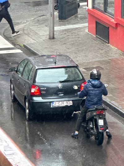 A person in a blue jacket and helmet is riding a motorcycle in front of a black Volkswagen Polo in Saint-Josse-ten-Noode, Belgium. The license plate on the car is 2-CJ-934. The motorcycle has a white license plate, but the number is illegible. The scene is wet, and the street is paved with large grey bricks. The person on the motorcycle is wearing white shoes. There is a silver pole to the left of the car with a sign that says "AO8." There are no pedestrians. The car is facing the camera, and the motorcycle is facing away from the camera. The weather appears to be rainy. The street is empty except for the car, the motorcycle, and the person on the motorcycle. The person on the motorcycle is the only person in the image. The image is taken from a high vantage point. The environment is urban. The location is identified using the coordinates 50.849, 4.378, which are the coordinates for Saint-Josse-ten-Noode, Belgium.