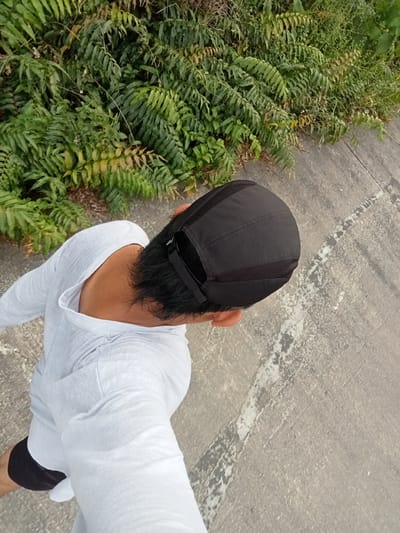 A person wearing a white long-sleeved shirt and black shorts is walking on a concrete path next to dense green foliage. The person is wearing a black cap and their back is to the camera, looking down at the ground. The concrete path has some white and grey markings and is slightly cracked. The foliage appears to be ferns and other plants, some of which are yellowing. The scene is outdoors and appears to be a quiet, possibly rural or suburban area, consistent with the location of Teluk Kenidai, Indonesia.