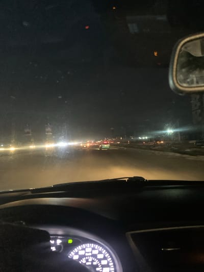 The image displays a night scene from the interior of a vehicle, looking out through the windshield onto a road. The perspective suggests a vantage point from the driver's or front passenger's seat. The primary image appears rotated 90 degrees clockwise.
Inside the vehicle, parts of the dashboard are visible on the right, featuring illuminated gauges with white numbers and markings, possibly a speedometer or tachometer. A green indicator light and other warning symbols, including a triangular hazard sign, are discernible on the dashboard. A rearview mirror is partially visible on the upper left, reflecting dark shapes within the car. A windshield wiper blade is also visible on the right edge of the frame.
The exterior scene is dark, illuminated by numerous artificial lights. Bright white streaks cut across the frame, indicating vehicle headlights or streetlights, while many smaller orange and red lights suggest vehicle taillights and other light sources, implying active traffic on the road. The road surface reflects these lights, which could be due to moisture or a dusty environment, contributing to the overall glare. Distant structures are faintly visible against the dark sky.
Notable visible text includes "3M" appearing in orange as a faint reflection or marking in the upper left quadrant of the full image. The scene captures the activity of driving or being transported in a vehicle at night on a populated street or highway. The location context is Jiru, Nigeria.