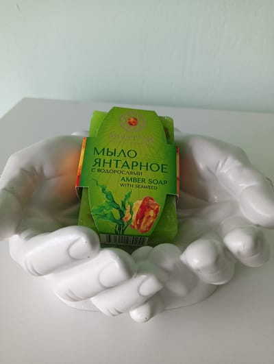 A green bar of amber soap with seaweed is presented in a white, stylized hand-shaped soap dish. The soap wrapper features a sun-like emblem and text in Russian, including "МЫЛО ЯНТАРНОЕ" (Amber Soap) and "С ВОДОРОСЛЯМИ" (with seaweed), as well as English text "AMBER SOAP WITH SEAWEED". The soap dish is resting on a white surface, likely a table or counter. The background is a plain, light blue wall. The scene is indoors, with neutral lighting. There are no people present. This image is located in Nizhnetroitskiy, Russia.