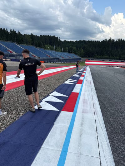 A brightly colored race track, adorned with blue, red, and white markings, stretches into the distance. The side of the track closest to the viewer has blue and white triangles, and the other side has red and white rectangles. A man wearing a black t-shirt and shorts stands on the colored track, while another man in an orange shirt is partially visible. Spectator seating lines the track beyond them, nestled near a dark green forest against a sky with fluffy white clouds.