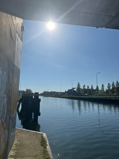 The image captures a bright, sunny scene along a canal in Weert, Netherlands, viewed from a slightly elevated position.
In the immediate foreground on the left, a large, imposing concrete structure, likely part of a bridge or embankment, dominates the frame. Its surface is a light grey, showing signs of weathering and some faint, light-colored graffiti patterns near its base. Atop this concrete structure, a small, dark grey box-like object with connected pipes or wires is visible.
The sun, intensely bright, is positioned in the upper left, casting a strong lens flare across the top portion of the foreground concrete and illuminating the clear blue sky.
The main subject is a calm canal or river that stretches horizontally across the mid-ground. Its surface is rippled, reflecting the clear sky and the distant green landscape. On the right side, partially submerged in the water and adjacent to the foreground structure, is a dark, substantial object, possibly a weathered wooden or metal component of a lock gate or a bridge support.
Along the distant right bank, a continuous line of green trees with varied foliage is visible, interspersed with several houses featuring dark roofs and multiple streetlights. A small blue and white sign can be seen among the trees on the far bank.
No specific human or animal activity is depicted; the scene is static and peaceful, bathed in bright daylight, suggesting a clear day in the morning or afternoon. The graffiti on the concrete wall is not clearly legible.