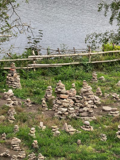 The image depicts a landscape in Staraya Ladoga, Russia, characterized by a body of water in the background, separated from the foreground by a rustic wooden fence. The foreground is a grassy slope dotted with numerous cairns, or stacks of stones. These cairns vary in size and composition, with some appearing to be carefully constructed and others more haphazard. The stones are predominantly gray and brown, reflecting the natural geological material of the area. The environment is verdant, with lush green grass and some interspersed dry vegetation. The overall scene suggests a natural, possibly historical or spiritual site, where visitors have engaged in the practice of stacking stones.