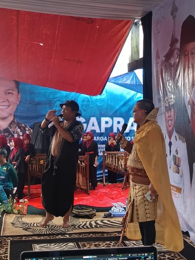 The scene depicts an outdoor event in Kawungluwuk, Indonesia, under a temporary canopy constructed from red and blue tarpaulins. Two men are central on a stage area covered with a zebra-patterned rug over a red tarp. The man on the left, barefoot, wears a dark bucket hat, an open patterned dark vest over a light garment, and dark loose pants, holding a microphone to his mouth while gesturing with his left hand. The man on the right, wearing sunglasses, is dressed in a gold and brown patterned traditional tunic, black pants, and a flowing gold cape, also holding a microphone to his mouth. Behind them, several women in dark attire and red headscarves stand next to wooden racks that appear to be angklung instruments; one woman holds a smartphone, seemingly recording. Large banners in the background feature portraits of individuals, including a man in a batik shirt on the left and a man in a white uniform adorned with medals on the right, with partial text "GAPRA" and "MULYADI" visible. A black bag and small white containers rest on the red tarp near the right man's feet. A laptop screen is partially visible in the bottom foreground.