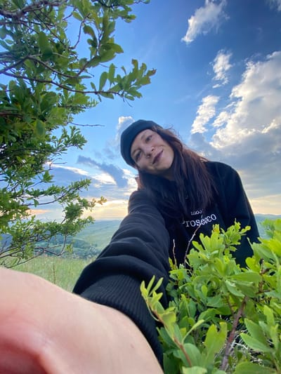 A medium shot captures a woman taking a selfie outdoors in a grassy area with a scenic landscape visible in the background. The woman is wearing a black beanie and a black sweatshirt with white text that reads "Prosecco." Her arm, also clad in black, extends towards the camera. She has dark hair, a nose ring, and is smiling. Lush green foliage frames the foreground on the left and right sides of the image. The sky above is blue with scattered white clouds. The background shows rolling hills, fields, and trees, suggesting a rural or natural setting. The location is identified as the city of Абзаново, Russia.