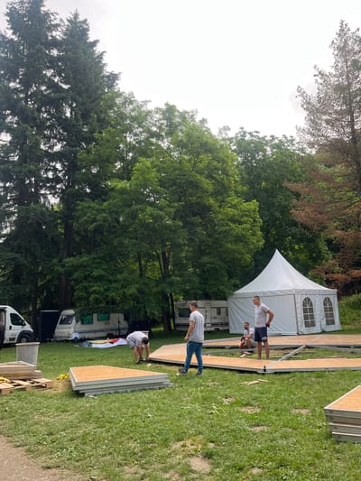 The image captures an outdoor scene in Sremski Karlovci Municipality, Serbia, depicting the setup for an event during daytime. Several men are actively engaged in assembling a temporary stage or flooring structure on a grassy field.
In the mid-ground, a large white event tent, featuring arched window panels, is set up. To the left of the tent and extending into the background, multiple camper vans or RVs are parked, alongside a white utility van in the foreground. The area where the men are working is covered with numerous wooden or plywood panels, laid out on metal framing to form a pathway or stage. More uninstalled panels are stacked nearby on the grass.
A man in a white t-shirt and dark shorts stands near the white tent, observing the progress. Closer to the foreground, another man in a grey t-shirt and blue jeans walks on a section of the constructed flooring. Further along the structure, a man in a white t-shirt kneels, appearing to work on the assembly.
The background is dominated by a dense treeline, suggesting a wooded or park-like environment. The sky above, visible in the upper left, appears overcast. The overall impression is one of active preparation for an outdoor gathering or festival. No discernible text is visible on any objects or signs in the image.