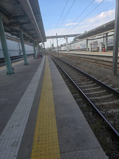The image shows an outdoor view of a train station platform in Agii Anargiri, Greece. The platform is paved with grey tiles and features a yellow tactile paving strip along the edge closest to the camera, followed by a white tactile strip. Several green pillars support a covered roof over the platform, which has advertisements and graffiti visible on the wall behind it. To the right of the platform, train tracks are laid on a ballast of grey stones, with some weeds growing between the sleepers. The tracks extend into the distance under a bright blue sky with scattered white clouds. In the background, there are overhead power lines and structures associated with the railway. A few people are seated on benches on the platform in the mid-ground, waiting.