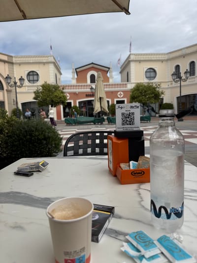 An outdoor cafe scene in Noventa di Piave, Italy, is captured from a first-person perspective at a table. The image shows a marble-patterned tabletop in the foreground, holding various items. A plastic water bottle filled with ice stands prominently, with a partial "RECO" or similar label visible. Next to it, an orange holder contains small blue sugar packets and a sachet dispenser labeled "bristot BELLUNO ITALIA 1919". A white stand with a QR code displays "Jody's Street Food Menù" and states "Non si effettua servizio al tavolo / No table service". An empty paper coffee cup with light brown residue is also on the table, along with a black pack of cigarettes, showing some Cyrillic text ("СИГАРЕТИ НАМАЛ"), and a black lighter.
The background reveals a bustling outdoor plaza with classic European architecture. Light-colored buildings with arched windows and prominent lamp posts line the square. One building has "ROSSY" inscribed on its facade. Green foliage from trees and bushes is scattered throughout the scene. Several Italian flags (red, white, green) fly from poles on the buildings. In the midground, blurred figures of people can be seen walking or sitting at outdoor tables with green umbrellas. The sky is overcast, indicating a cloudy daytime setting. The overall atmosphere suggests a casual moment at a cafe in a public square.