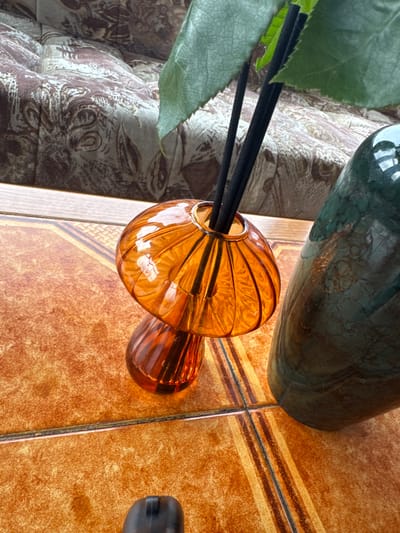 Inside a residence in Fowlis Easter, United Kingdom, a close-up, elevated view captures three primary objects on a mottled orange-brown tiled surface. Centrally positioned is an orange, mushroom-shaped glass diffuser bottle featuring vertical fluting on both its cap and stem, containing several slender black diffuser reeds. Partially obscuring the reeds and extending from above is a single, vibrant green plant leaf. To the right, a tall, dark green bottle or vase with a subtle marbled texture stands. A small, dark, indistinct object is partially visible in the lower foreground on the tiled surface. The tiled surface exhibits square formations with decorative brown and yellow striped patterns. In the background, separated by a light-colored wooden strip, a beige and brown upholstered surface with a prominent abstract or floral pattern is visible, likely a sofa or chair.