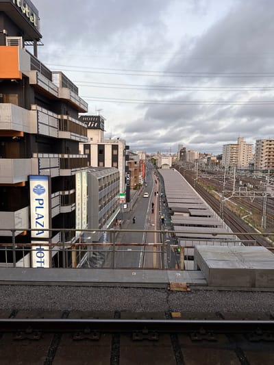 This is a medium-angle, eye-level shot of a street and train tracks in Funabashi, Japan. The scene is dominated by a multi-story building with dark grey and white tiled facades, and an orange accent wall on the left side of the image. A large blue and white sign reads "PLAZA HOTEL" vertically on the left building. Other buildings, including what appear to be hotels, line the street. The street itself is paved, with sidewalks and streetlights. Several people are visible walking on the sidewalk. To the right of the street are train tracks with a covered platform. The sky is overcast with dramatic clouds. The foreground features train tracks, ballast, and a metal barrier, suggesting the photo was taken from a train platform or an overpass.