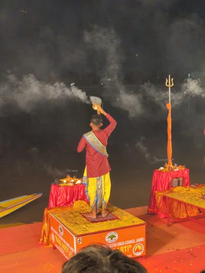The image depicts a spiritual ritual, likely a religious ceremony, taking place at night, possibly on the banks of the Tawi River in Jammu, India, given the presence of the "Maha Aarti" inscription which is a well-known event in Jammu. A young man, dressed in traditional attire consisting of a maroon kurta and a yellow dhoti, stands on a raised platform adorned with yellow flowers. He is performing a 'puja' or ritual, holding a brass vessel with smoke emanating from it, indicating the burning of incense or offerings.
In the background, a tall, golden trident, a symbol associated with Lord Shiva, stands prominently. Red draped tables are visible, one holding offerings like flowers, shells, and lamps, while another table to the right is also laden with yellow flowers. Smoke or mist hangs in the dark, hazy atmosphere, obscuring distant lights, suggesting an outdoor setting perhaps with a fog machine or natural atmospheric conditions. The overall scene is illuminated by artificial lights, casting a warm glow on the ritualistic elements. The presence of a boat in the lower left corner further hints at a riverside location. The platform the man is standing on is an orange box with inscriptions that read "SHRI RAM BHAKTA GANESH YOG MANDAL" and "MAHA AARTI" followed by "RIDOR VIKAS COUNCIL JAMMU".