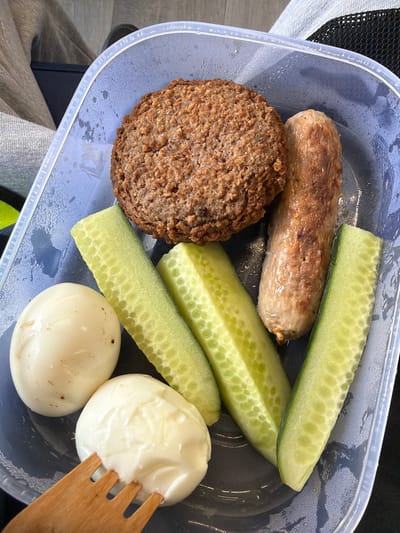 A top-down shot shows a partially filled, rectangular, transparent plastic container with a black interior base. The container holds a selection of food items: a browned, circular meat patty; a cooked sausage with a light brown casing; four sticks of cucumber, uniformly cut; and two peeled hard-boiled eggs, one with a dollop of white creamy sauce. A wooden fork is partially visible, resting against one of the eggs coated in sauce. The background reveals details of what appears to be clothing or fabric, with the edge of a bag visible at the top left and green/yellow stripe. No information to locate the image within Connah's Quay, UK is contained within the image.