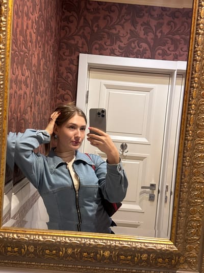 A young woman with light brown hair pulled back is reflected in an ornate gold-trimmed mirror, taking a selfie. She is wearing a light blue denim jacket over a cream-colored turtleneck. Her left hand is raised to her head, touching her hair. She is looking at the phone in her right hand. Behind her, a cream-colored door with raised panels is visible. The wall to her left is covered in patterned maroon wallpaper. The scene is indoors, likely in a bathroom or entryway, and the location is not identifiable as Shkotovo, Russia.