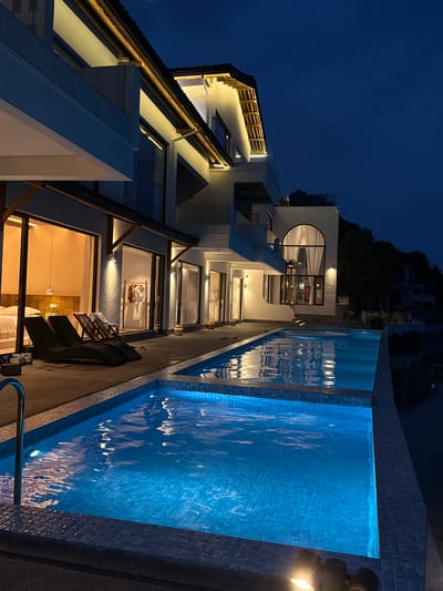 The image depicts a modern building complex adjacent to a swimming pool at night. The complex features a multi-story structure with white exterior walls and a combination of large windows and balconies. The balconies and windows are illuminated, revealing interior spaces with furniture. One distinctive architectural feature is a large, rounded window on one of the upper floors. A dark rooftop is visible at the edge of the structure.
Next to the building, a swimming pool is filled with illuminated blue water, reflecting the surrounding light. The pool is bordered by dark tiles.
The sky is a deep blue, indicating it is nighttime. The scene is lit by artificial lights, highlighting the building and pool, creating a vibrant and contemporary atmosphere.
No people are visible in the frame. The image focuses on the architectural design and the ambience of the location. Jinhua, China is specified as the location.