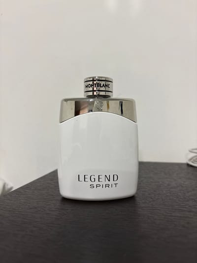 The image displays a Montblanc Legend Spirit perfume bottle centered on a dark brown, flat surface. The bottle is opaque white with "LEGEND" printed above "SPIRIT" in black capital letters on its front. It features a silver-toned metallic cap, which has "MONTBLANC" embossed in black capital letters on its top circular edge and a visible Montblanc star emblem on its front face. The background consists of a plain, light-colored wall. A white, coiled object, possibly a cable or fabric, is partially visible on the dark surface towards the upper right side of the frame. No individuals are present, and no actions or interactions are depicted. The scene is asserted to be located in the city of Quimbaya, Colombia.