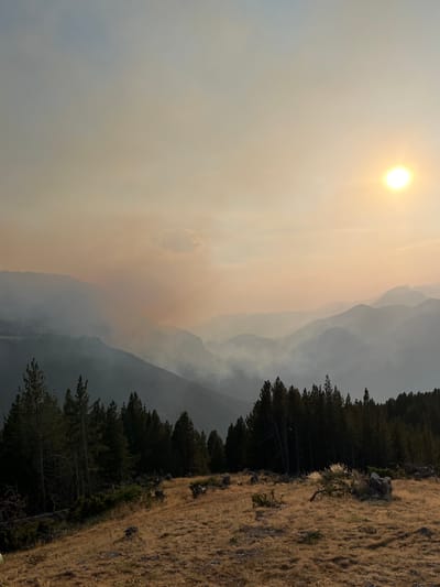 An outdoor, elevated, wide shot captures a landscape dominated by hazy mountain ranges under a sun partially obscured by atmospheric smoke. The foreground features a grassy slope dotted with sparse trees and rocky outcrops. A dense line of pine trees forms a dark band across the middle ground, with more trees visible on the slopes of the mountains in the distance. The sky is a muted orange and yellow, indicative of smoke, with the sun appearing as a bright white circle in the upper right quadrant. The overall impression is one of natural beauty impacted by environmental conditions, potentially a wildfire, contributing to the hazy atmosphere and the muted sunlight. The location is identified as San Juan de Plan/San Chuan de Plan, Spain. There are no people or animals readily visible in the image.
