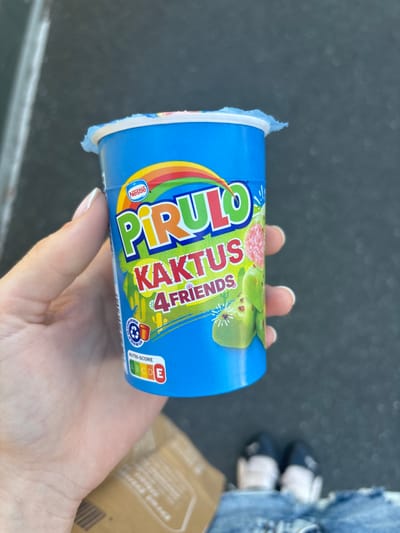 A person's hand is holding a blue yogurt cup. The cup has a colorful label with the text "PIRULO KAKTUS 4 FRIENDS" printed in large, bold letters. The brand "Nestlé" is visible in the bottom left corner of the label. There are illustrations of cactus and green fruits on the label. The hand holding the cup has light skin and manicured fingernails. In the background, a person wearing blue jeans and white and navy shoes is visible, and a brown paper bag. The setting appears to be outdoors on a paved surface. The image is well-lit, suggesting it was taken during the daytime. The context of Erlenbach am Main, Germany, provides a geographical location. No specific weather conditions or emotions are discernible from the image. The Nutri-score rating of A, B, C, D, E is displayed on the right side of the yogurt cup label.